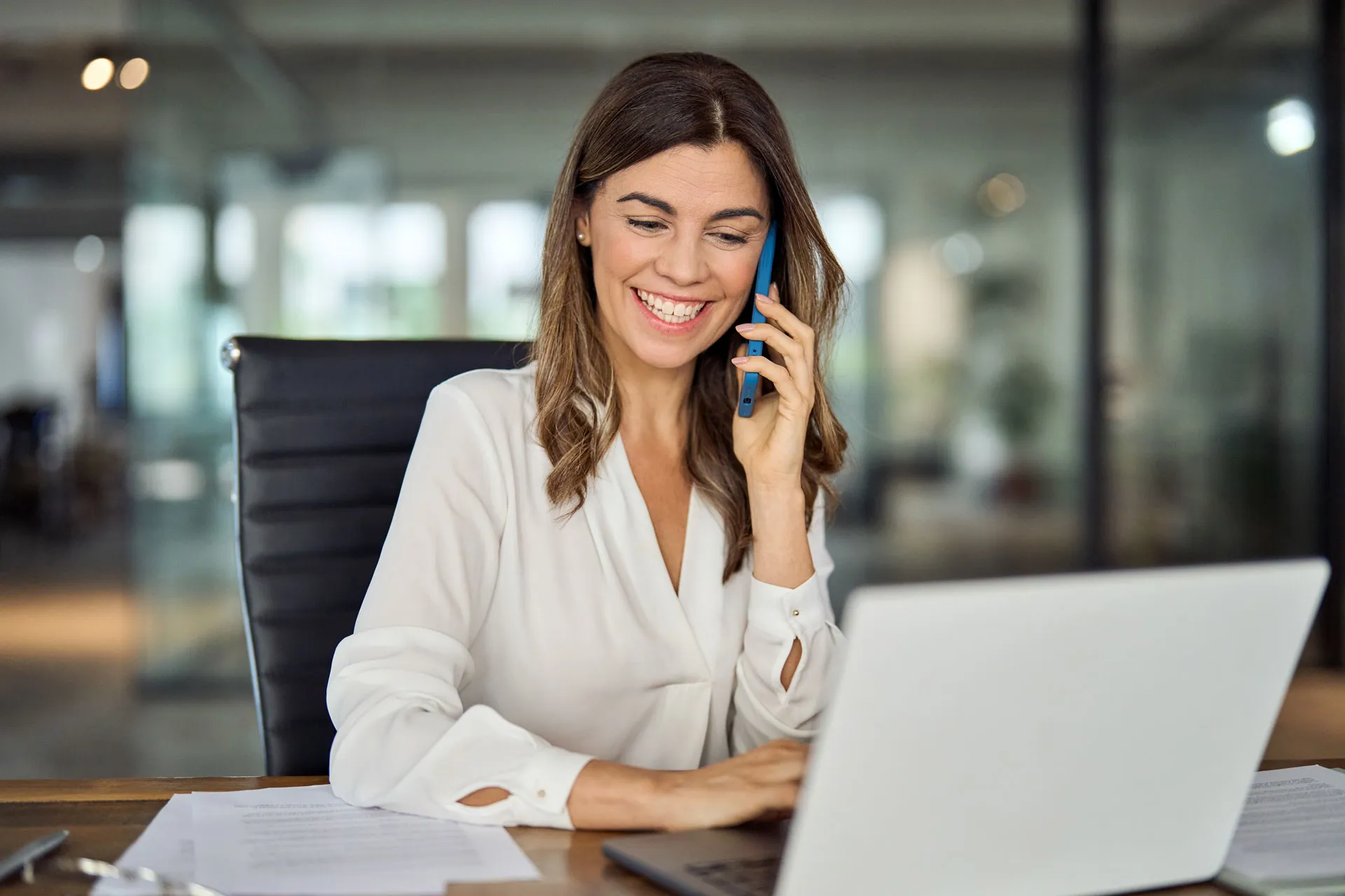 Lächelnde Frau sitzt an ihrem Arbeitsplatz, telefoniert und arbeitet am Laptop